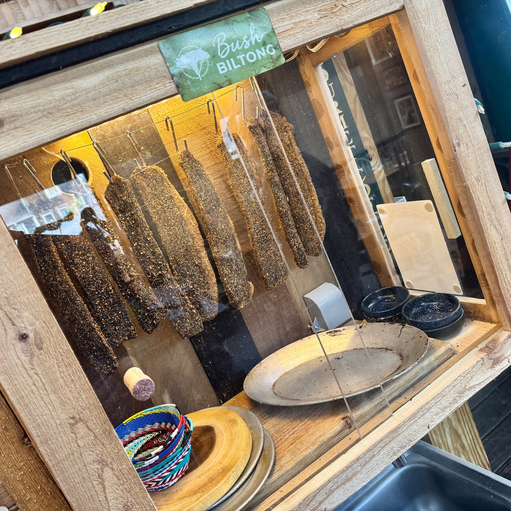 Dried meat products hanging in a wooden display case with a 'Bush Biltong' sign.