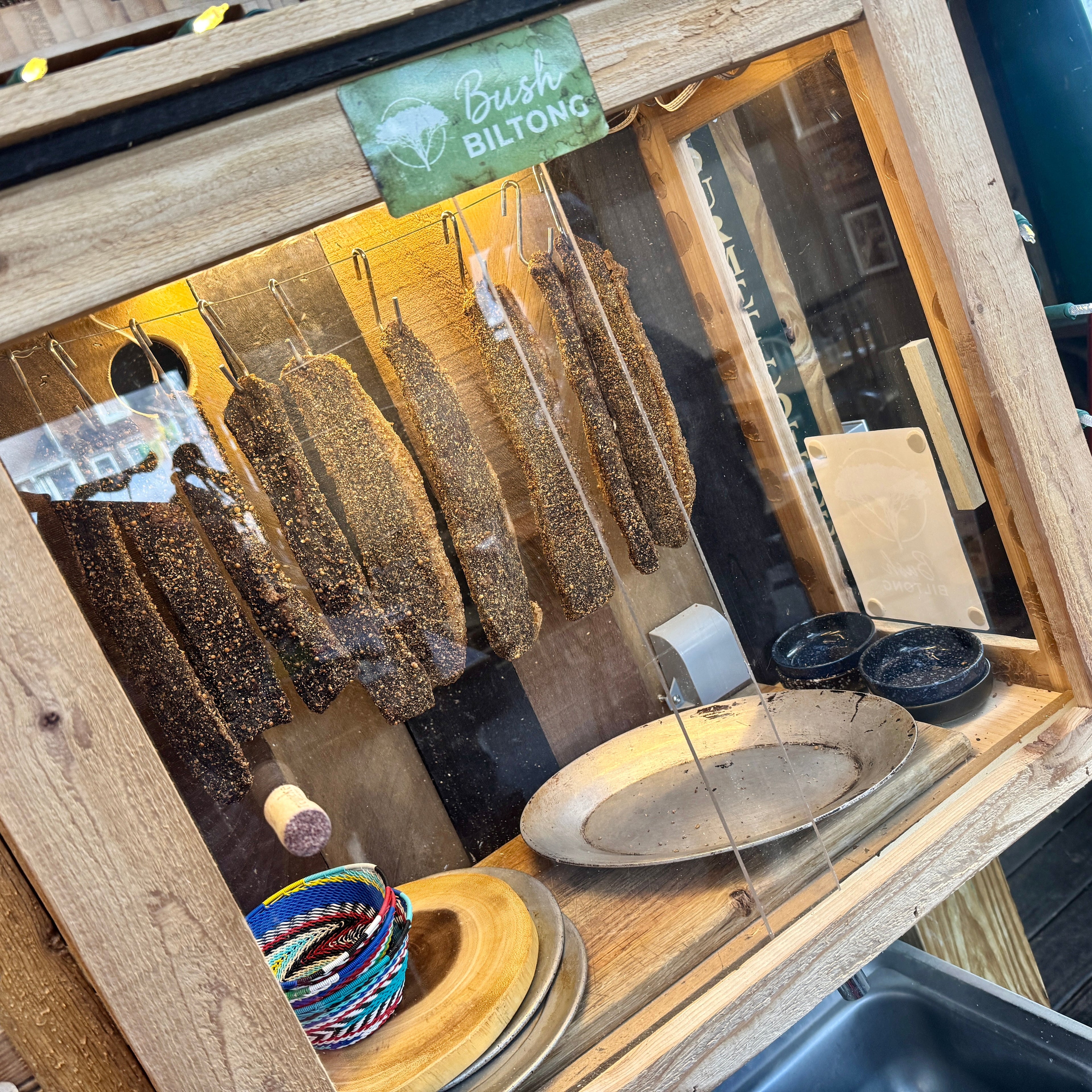 Dried meat products hanging in a wooden display case with a 'Bush Biltong' sign.